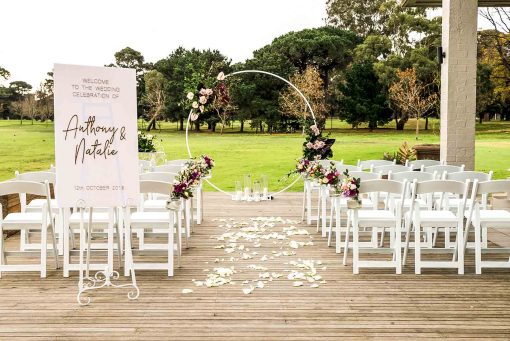 Contemporary wedding ceremony setting showing chairs, white circular arch with floral arrangements and signage