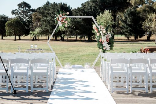 White Hexagon Arch with florals, white wedding chairs with a green leafy backdrop