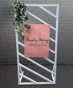 Blush pink plinth and cake stand in front of white window panel backdrop 2m high by 1m wide with floral adornment and blush pink sign attached that says ‘Simply Staging Property Styling’.