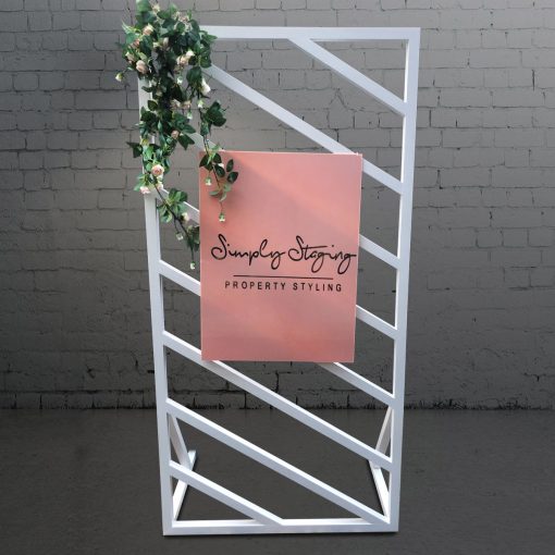 Blush pink plinth and cake stand in front of white window panel backdrop 2m high by 1m wide with floral adornment and blush pink sign attached that says ‘Simply Staging Property Styling’.